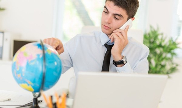 Businessman using phone and looking at globe at discovering available markets of global business. He pointing finger on the globe on the table in front of him.